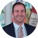 Professional headshot of smiling orthodontist in suit and tie against blurred dental office background